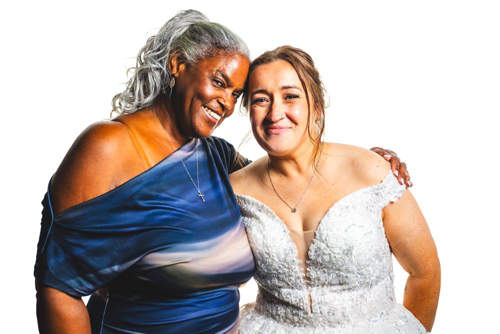Two women smiling together, one in a blue dress and one in a white lace wedding gown, posing for a bridal photo.