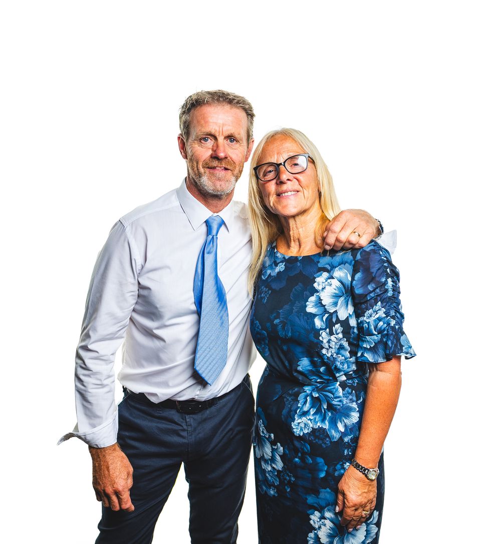 A smiling man in a blue shirt and tie poses with a woman in a floral blue dress against a white background.