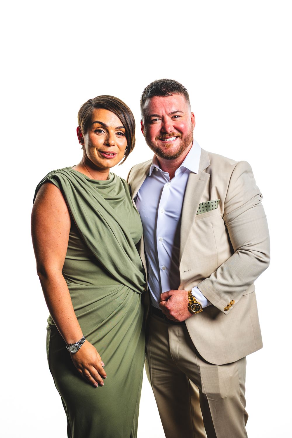 A smiling couple poses together, woman in green dress and man in beige suit, against a white background.