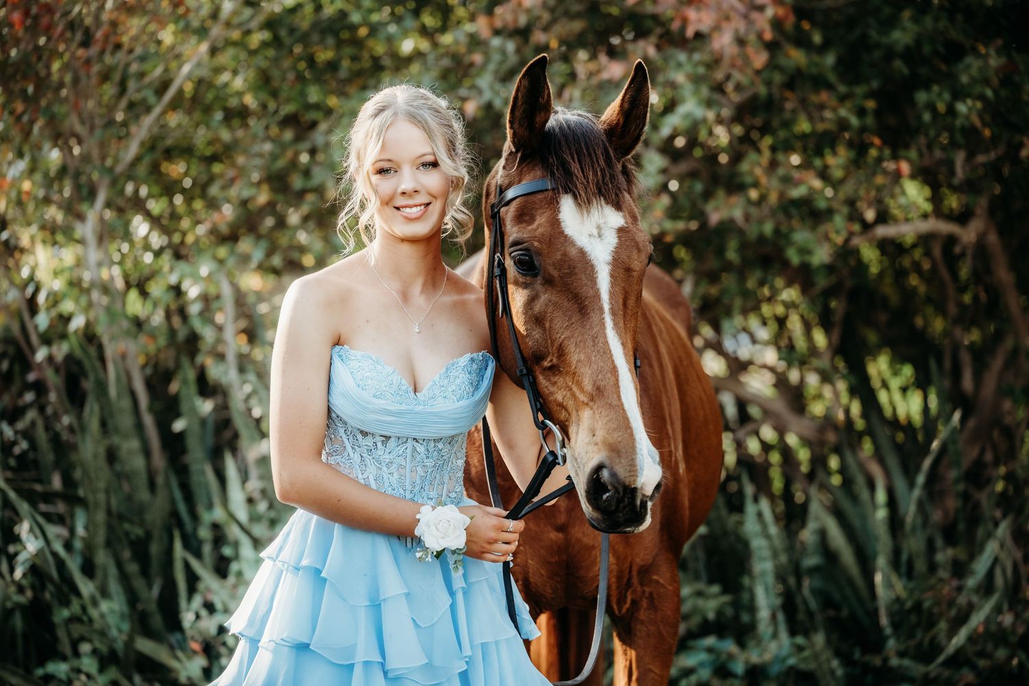 Young woman in blue strapless ball gown smiles beside a chestnut horse outdoors among lush green foliage.
