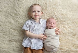 Smiling toddler boy lying on fluffy rug with sleeping swaddled newborn sibling in soft natural light.