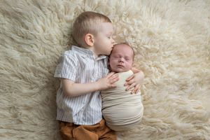 A toddler boy gently kisses a swaddled newborn baby lying on a soft cream fur rug.