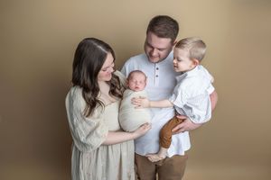 Family of four posing in studio, parents holding newborn baby and toddler son against warm beige background.