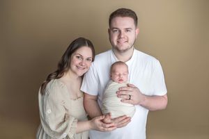 Young couple smiling and holding their swaddled newborn baby against a warm tan studio background.