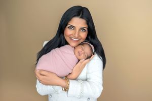 A smiling woman with long dark hair holds a swaddled newborn baby in a pink wrap during a studio portrait session.