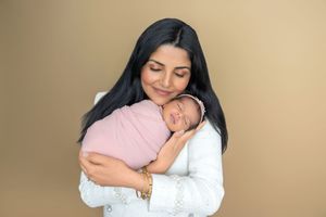 A mother smiles warmly while cradling her sleeping newborn wrapped in a pink swaddle against a tan background.