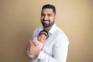 Smiling bearded man in white shirt holding sleeping newborn baby wrapped in pink swaddle against beige background.