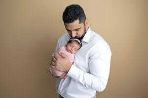 A father in a white shirt tenderly holds his swaddled newborn baby against a warm beige background.