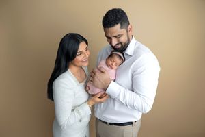 Couple smiling and holding their newborn baby wrapped in pink against a warm beige studio background.