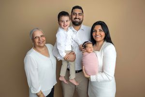 Multi-generational family portrait with grandmother, parents, toddler son, and newborn baby posing against beige background.