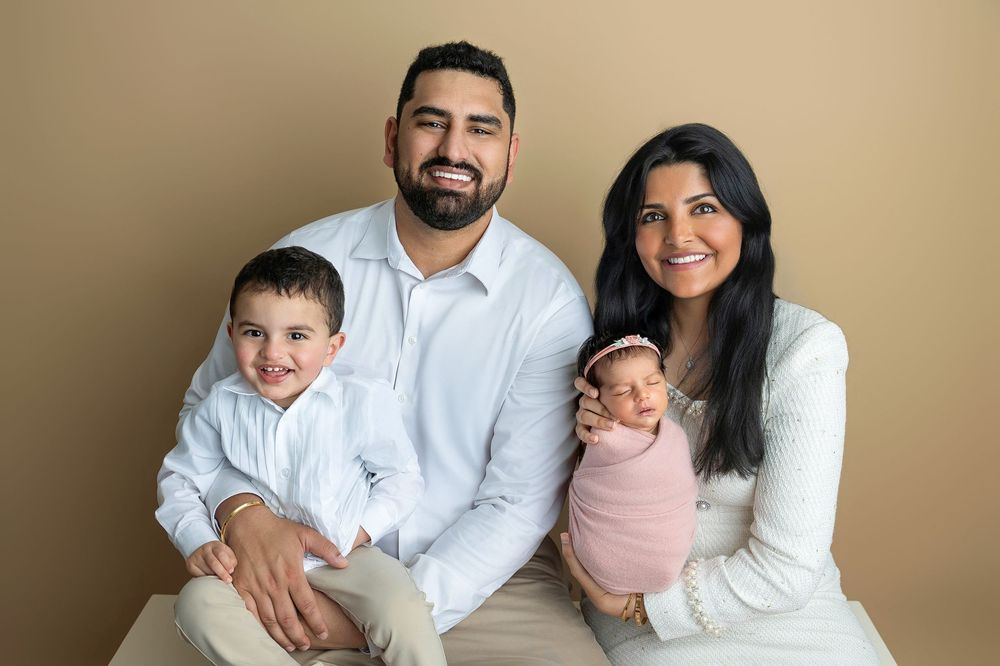 Family portrait with parents and two young children, including a newborn in pink wrap, posed against a warm beige background.