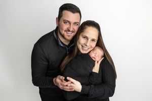 A smiling couple in black outfits holds their sleeping newborn baby against a white studio background.