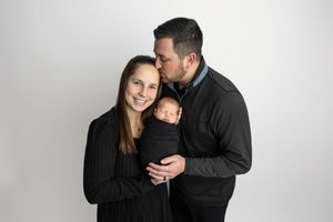 A smiling couple in black clothing holds their swaddled newborn baby while the father kisses the mother's head.