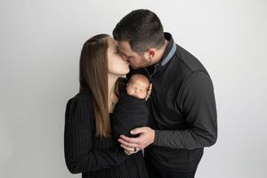 New parents kissing while tenderly holding their swaddled newborn baby in a professional studio portrait.