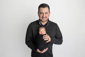 Smiling man in black sweater holding a sleeping newborn baby wrapped in dark swaddle against white background.