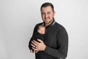 Smiling man in dark sweater holding sleeping newborn baby wrapped in black swaddle against white background.