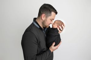 A father tenderly kisses his swaddled newborn baby while holding them close against a white background.