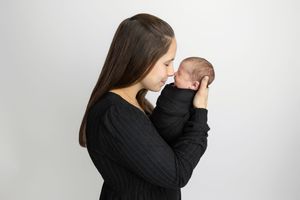 Young mother in black sweater tenderly holds and kisses her newborn baby against a white background.