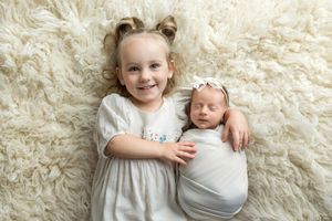Young girl with pigtails smiling beside swaddled newborn baby sister on fluffy white rug.