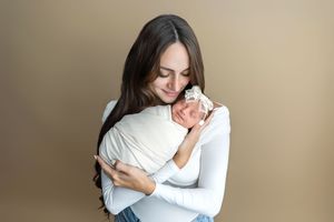 A mother tenderly holds her sleeping newborn baby wrapped in white against a neutral beige background.