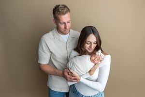 A couple tenderly cradles their newborn baby wrapped in white, posing together against a neutral beige background.