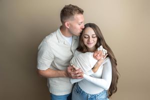 A couple embraces tenderly, man kissing woman's head, both smiling softly against a neutral beige background.