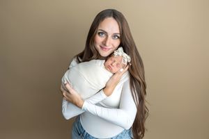 A smiling woman with long brown hair holds a sleeping newborn baby wrapped in white against a tan background.