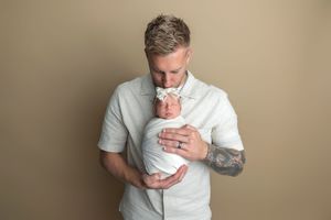 Father with tattoos tenderly kisses swaddled newborn baby in white wrap with bow in professional studio portrait.