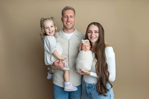 Happy family of four posing for a newborn portrait, parents holding toddler girl and swaddled baby on beige background.