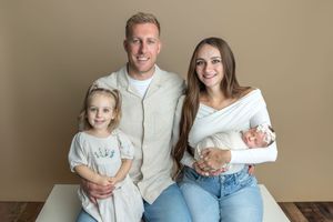 Family portrait of parents with toddler girl and newborn baby posing together in a professional studio setting.