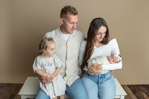 Family of four posing for newborn portrait, parents seated with toddler girl and swaddled baby in neutral studio setting.