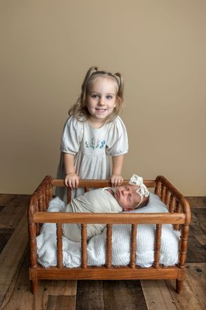 A smiling young girl with pigtails stands behind a wooden doll bed holding a swaddled newborn baby wearing a white bow headband.