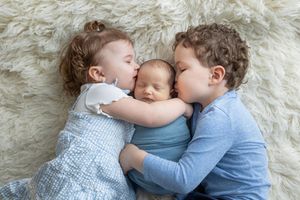 Two young siblings kissing a sleeping newborn baby wrapped in blue on a fluffy white blanket.