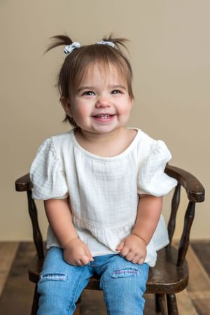 Smiling toddler girl with pigtails wearing white top and jeans sitting on wooden chair.