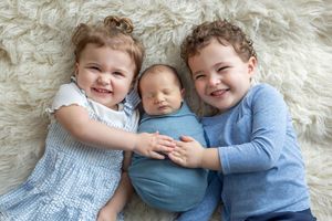 Two smiling toddlers in blue outfits lying on fluffy rug with newborn baby swaddled in teal between them.