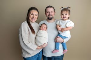 Happy family of four posing for newborn portrait, parents holding swaddled baby and toddler girl against tan background.