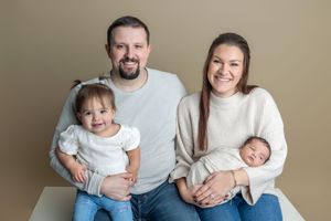 Family portrait of parents with toddler girl and newborn baby, smiling against a neutral beige background.