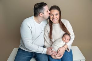 Happy couple in white sweaters sit together smiling, father kissing mother on cheek while she holds sleeping swaddled newborn.
