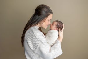 Mother tenderly holding swaddled newborn baby close to her face against olive background.