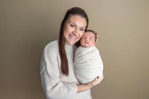 Smiling mother in white sweater holds sleeping swaddled newborn baby with headband against neutral background.