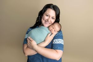 Smiling mother holding sleeping newborn baby wrapped in teal swaddle against beige studio background.