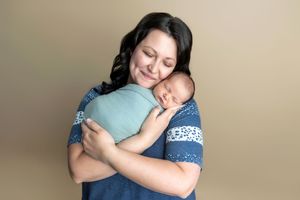 A mother smiles warmly while cradling her swaddled newborn baby against her chest in a studio portrait.