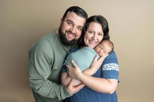 Smiling parents pose with their swaddled newborn baby against a neutral tan background in a professional portrait.