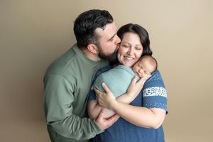 Father kisses mother on cheek as she smiles holding swaddled newborn in a warm studio family photo.