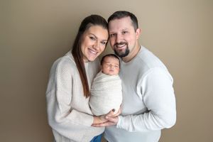 Smiling parents holding their swaddled newborn baby against a neutral tan background in a professional portrait.