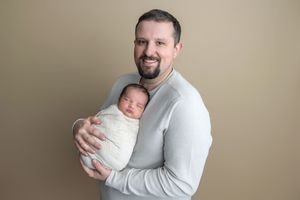 A smiling father in a gray shirt holds his swaddled newborn baby against a neutral tan studio background.