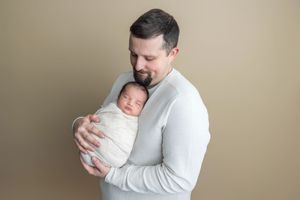 A father tenderly holds his swaddled newborn baby close to his chest against a neutral beige studio background.