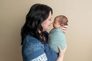 A mother with dark hair holds her swaddled newborn baby close, sharing a tender moment against a neutral background.