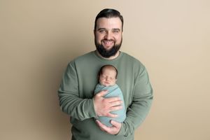 Smiling bearded man in green sweatshirt holding a sleeping newborn baby wrapped in blue, studio portrait.