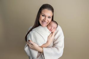 A smiling mother with dark hair holds her swaddled newborn baby close to her chest in a professional studio portrait.